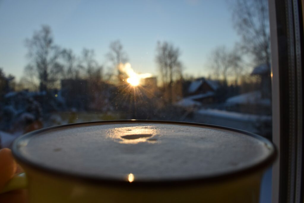 A cup of coffee on a windowsill with the winter sun rising behind bare trees